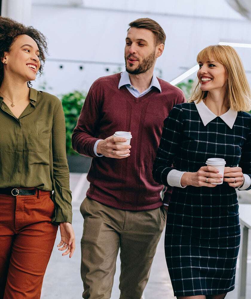 Three diverse colleagues walking together holding coffee cups in bright modern office space