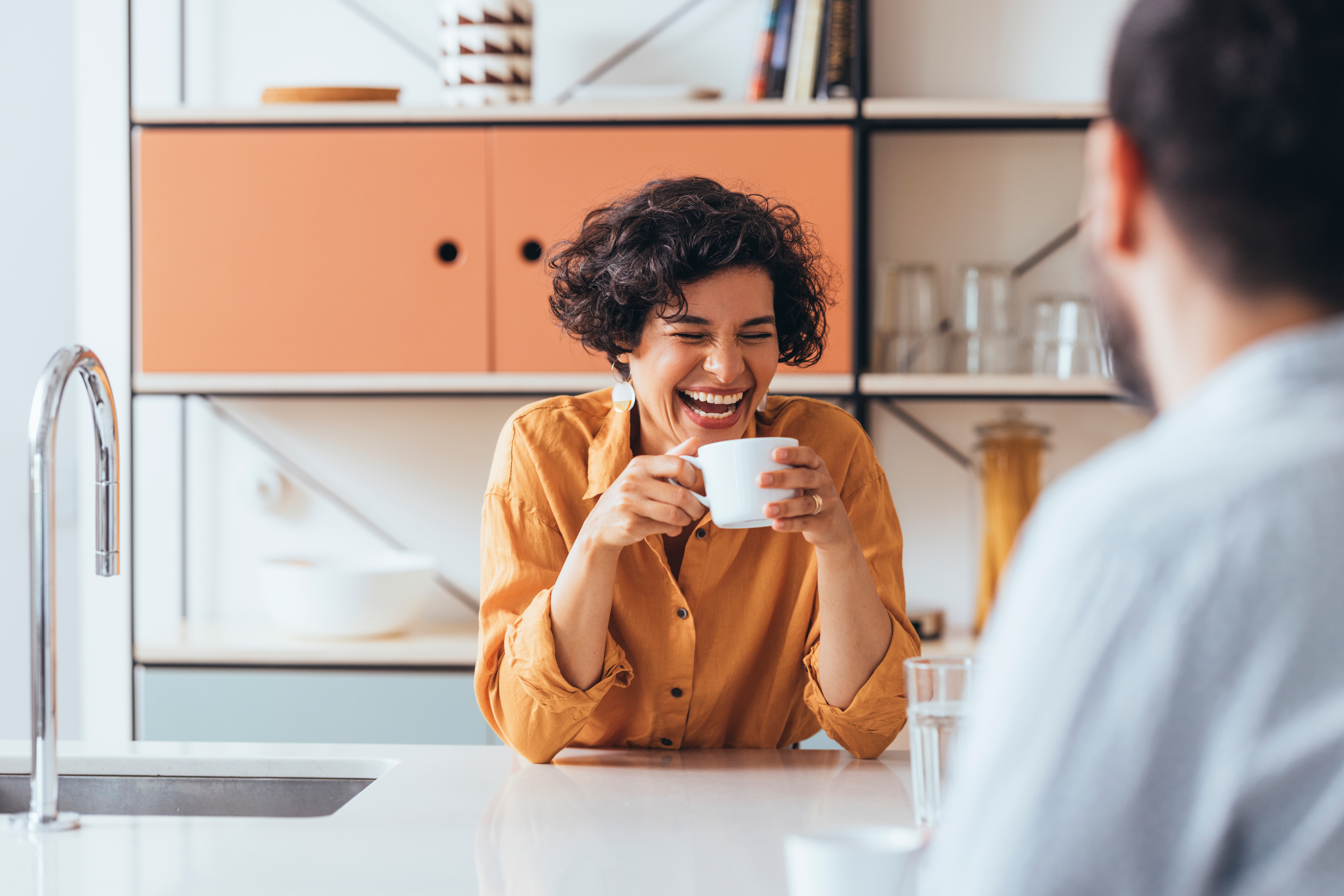 Woman with curly hair laughing while holding white coffee cup in modern kitchen with orange cabinets